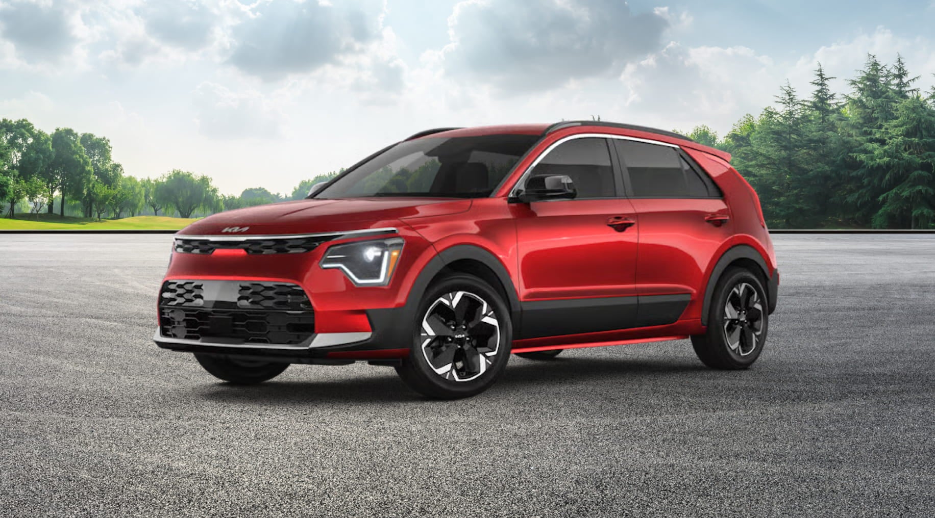 A red Kia Niro EV is parked on a paved surface with a backdrop of green trees and a cloudy sky.